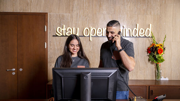 Zwei fröhliche Mitarbeiter:innen stehen an der Rezeption von magdas HOTEL. An der Wand ist ein Schriftzug mit den Worten &quot;stay open-minded&quot; zu sehen.